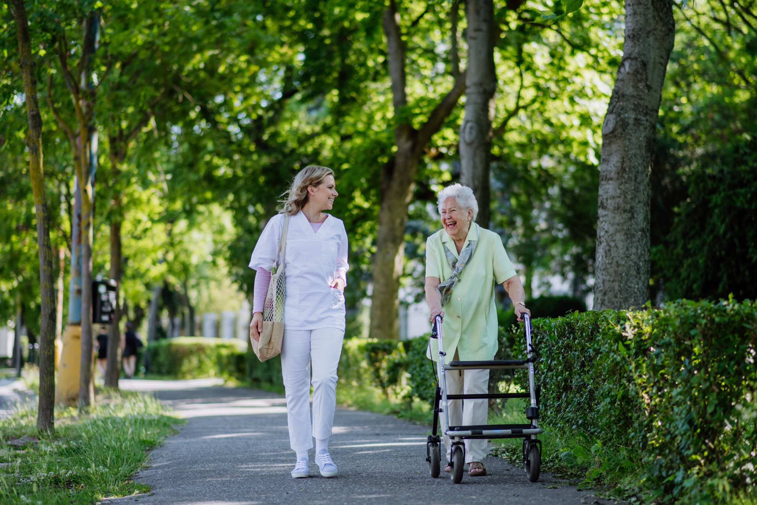 A younger and older woman walking and smiling together. The older woman is using a walker.