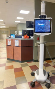 A photo of the telestroke robot in the corner of the Harney District Hospital Emergency Department. The robot consists of a computer screen and webcam with zoom lenses that is mounted on a tall stand with wheels. 